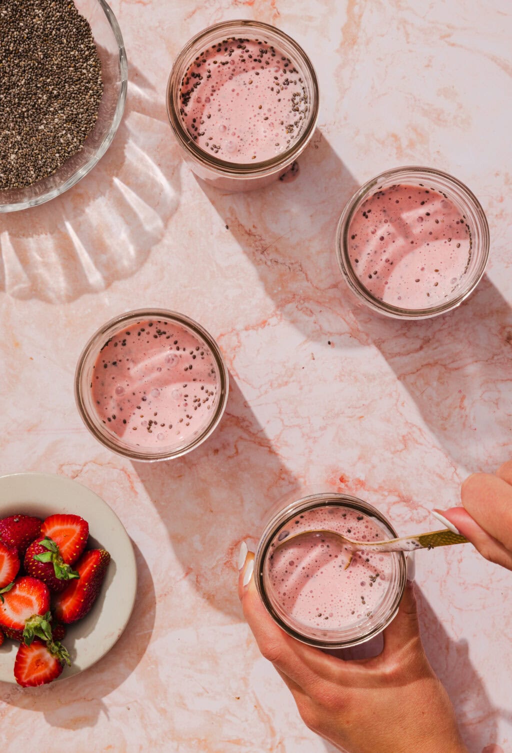Four glasses of pink chia seed pudding on a marble surface, with a small bowl of chia seeds and a plate of sliced strawberries nearby. A hand stirs one glass with a spoon.