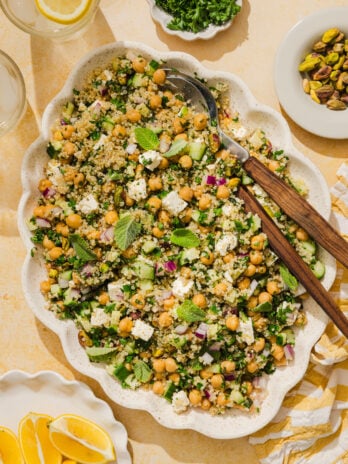 A platter of chickpea and quinoa salad with feta, red onion, cucumber, fresh herbs, and mint leaves, surrounded by lemon wedges, pistachios, chopped parsley, and glasses of water.