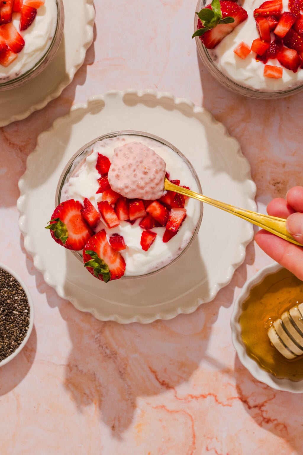 A hand holds a spoon over a bowl of yogurt topped with chopped strawberries. Two similar bowls, chia seeds, and honey with a honey dipper are nearby, all on a light marble surface.
