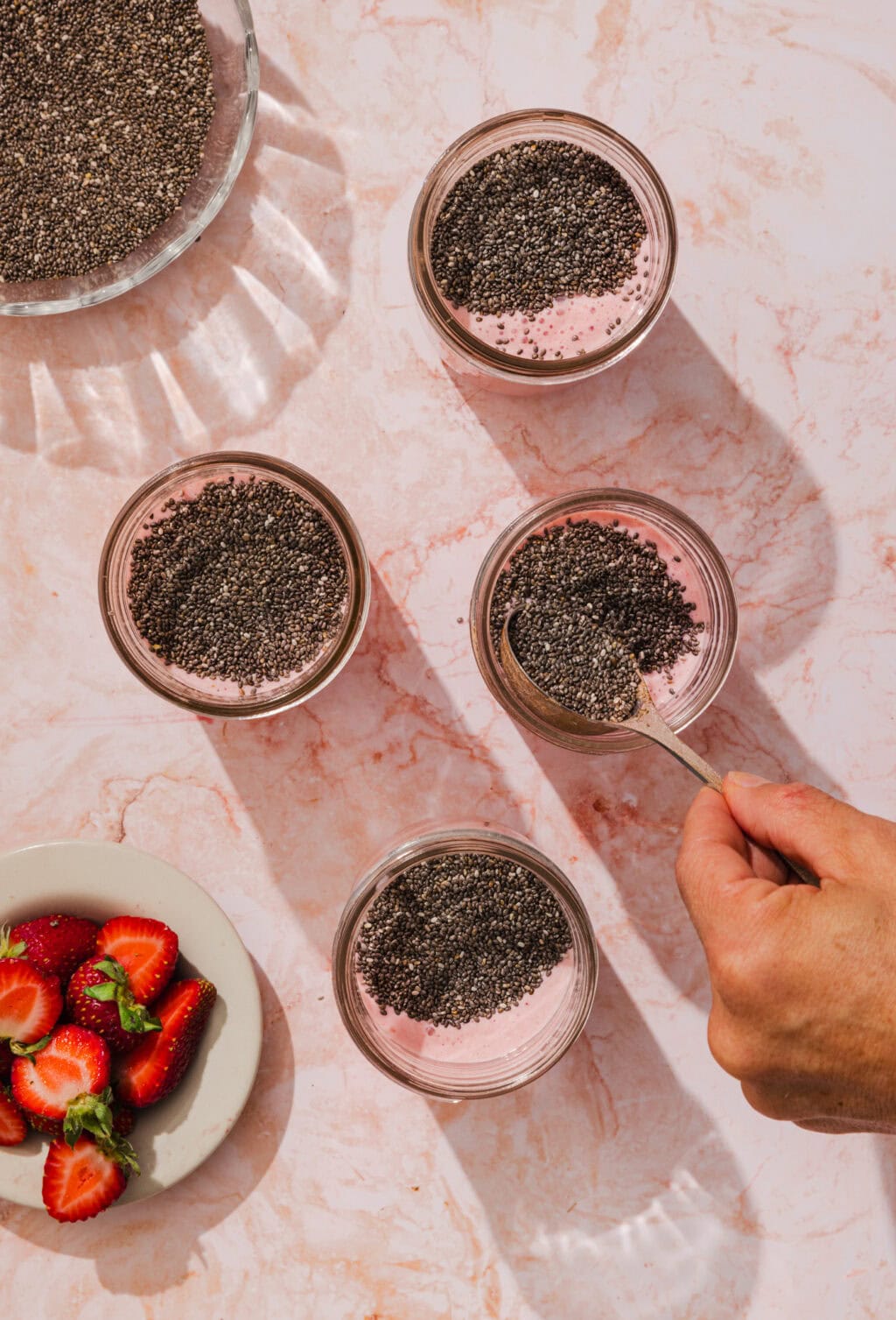 Four small glass containers filled with pink yogurt or smoothie and topped with chia seeds. A hand holds a spoon over one jar. A small plate of sliced strawberries is nearby on a marble surface.