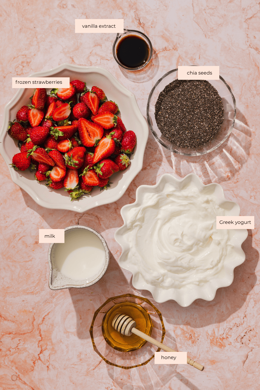 Bowls of frozen strawberries, chia seeds, Greek yogurt, a small pitcher of milk, a dish of honey with a honey dipper, and a small bowl of vanilla extract are arranged on a pink marble surface—perfect for preparing strawberry chia pudding.