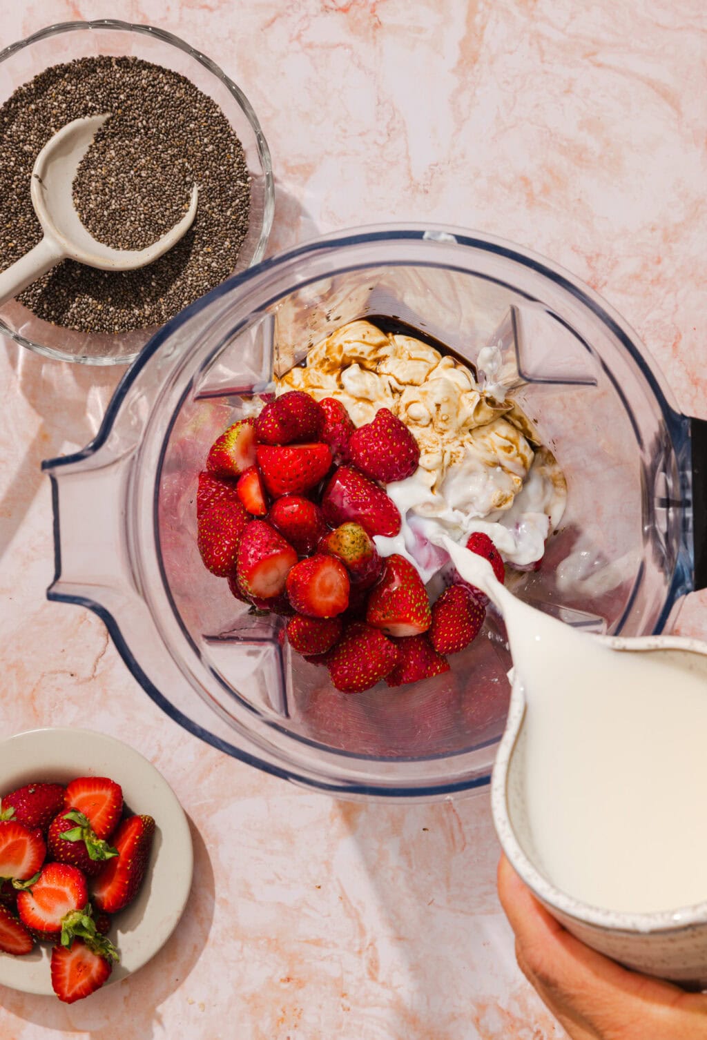 A blender contains yogurt, oats, and strawberries while milk is being poured in. Nearby, theres a bowl of chia seeds with a spoon and a plate of sliced strawberries on a pink marble surface.