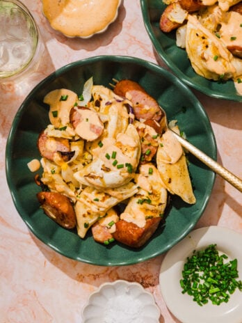 A green bowl with pierogi, sliced sausage, onions, creamy sauce, and chopped chives on top, with a fork. Surrounding dishes hold chives, salt, extra sauce, and a glass of water on a pink marble surface.