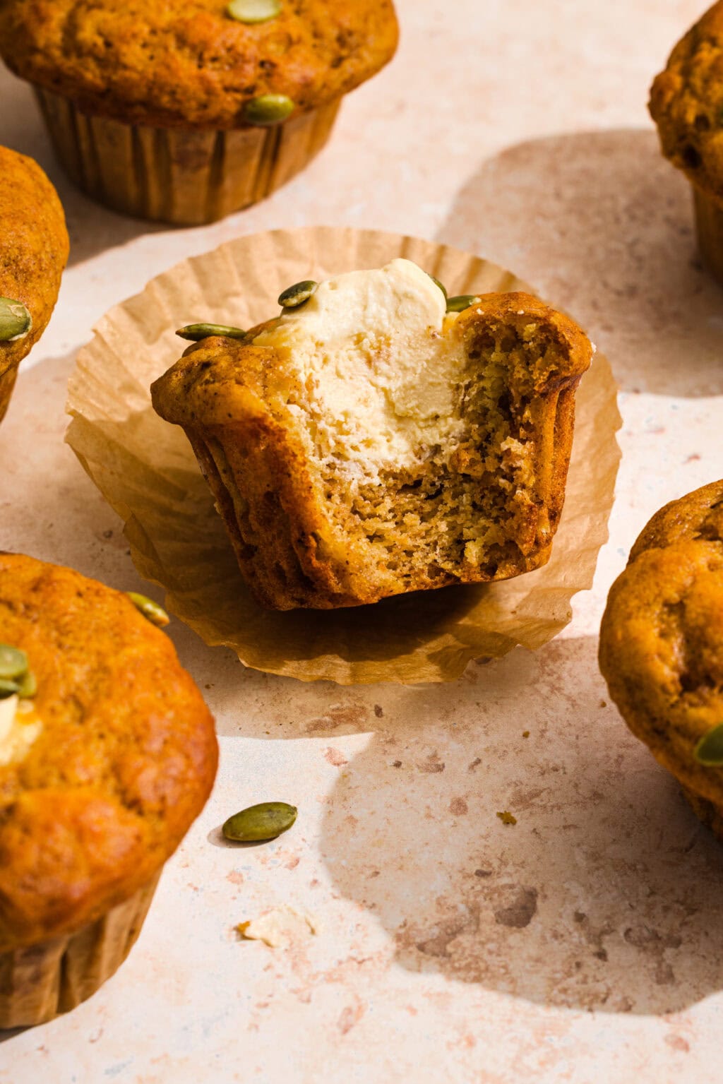 A pumpkin muffin with cream cheese filling, partially eaten, sits on a parchment wrapper. Whole pumpkin muffins and scattered seeds are nearby on a light textured surface.
