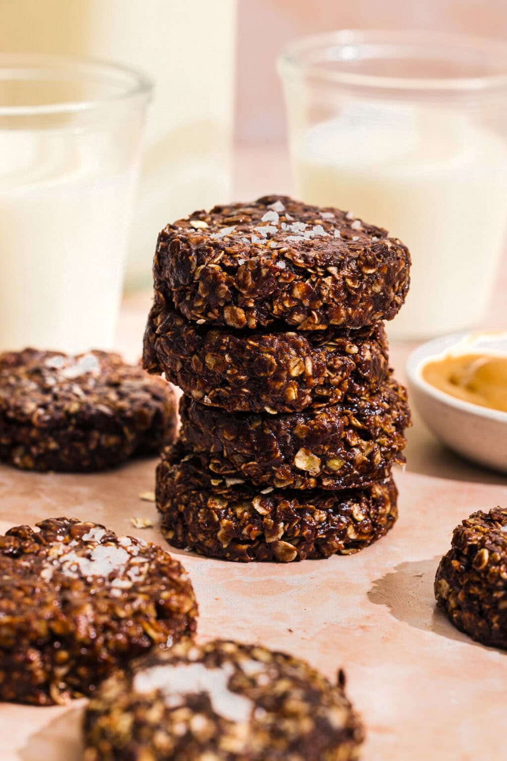 A stack of homemade chocolate oatmeal cookies sits on a light surface, surrounded by more cookies, a bowl of peanut butter, and glasses of milk in the background.