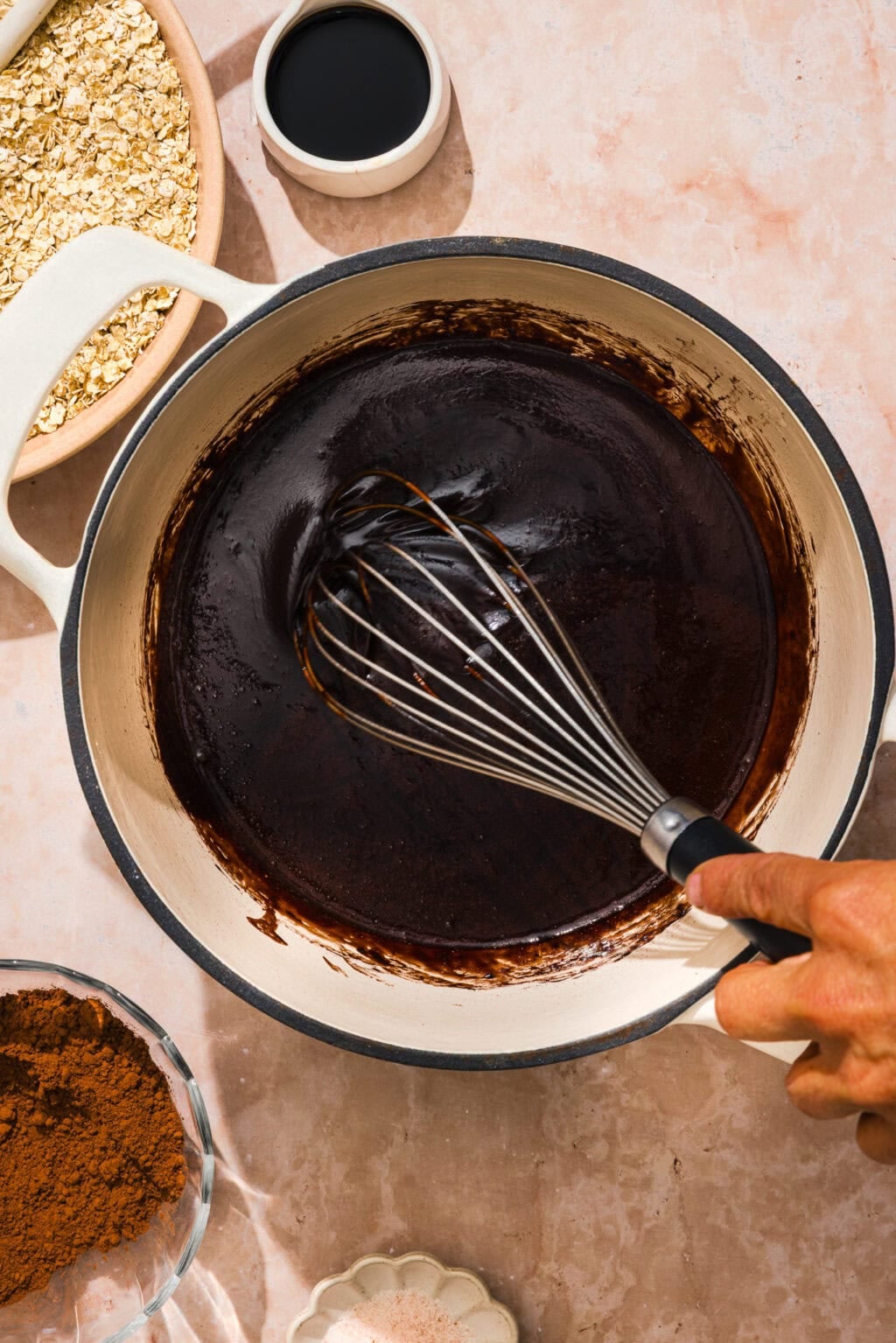 A hand stirs glossy melted chocolate in a white pot with a whisk. Surrounding the pot are bowls of oats, cocoa powder, and a small bowl of dark liquid, all on a light-colored countertop.