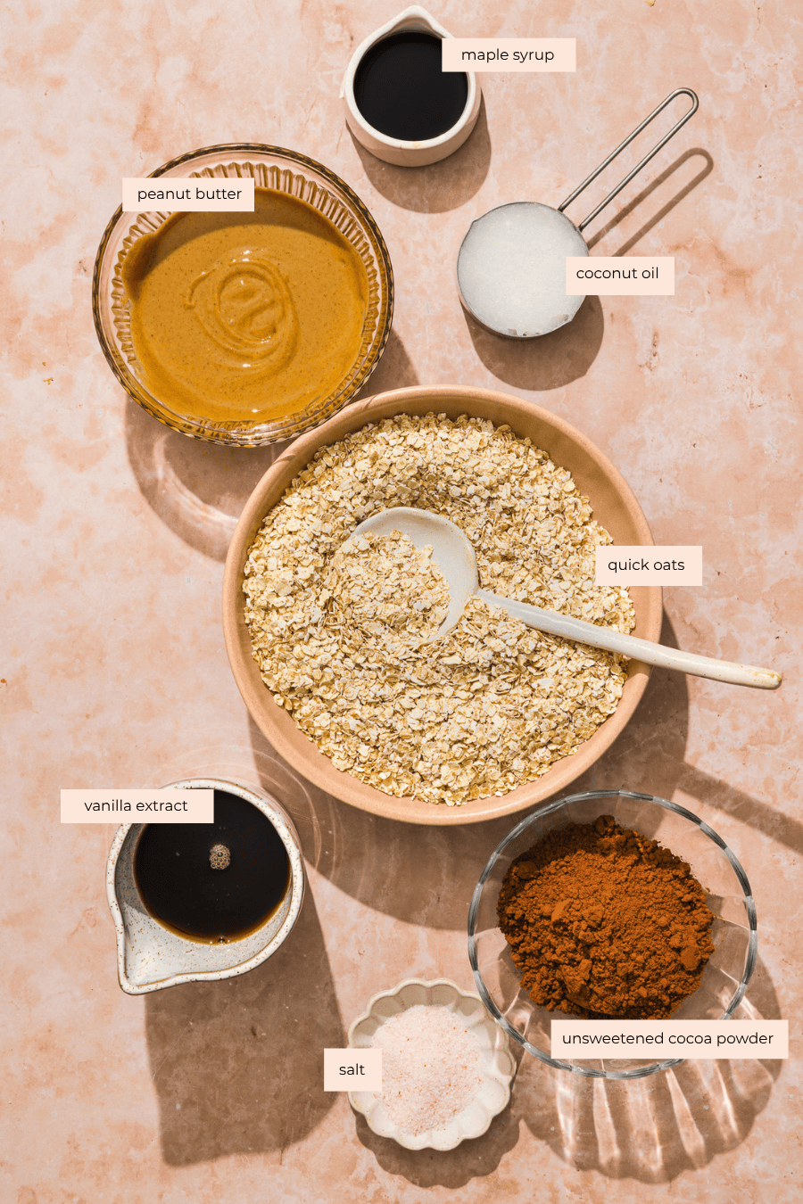 Bowls and measuring cups with labeled ingredients: maple syrup, peanut butter, coconut oil, quick oats, vanilla extract, unsweetened cocoa powder, and salt, arranged on a light pink surface.