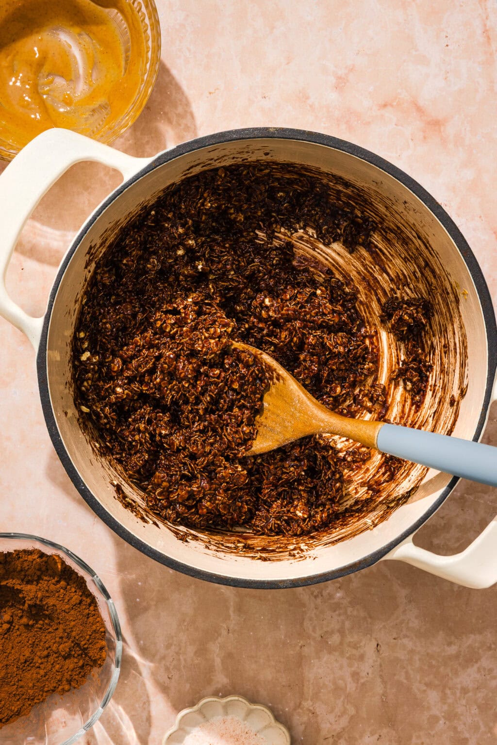 A pot filled with a chocolate oat mixture being stirred with a wooden spoon, surrounded by bowls containing cocoa powder and a creamy ingredient on a light countertop.