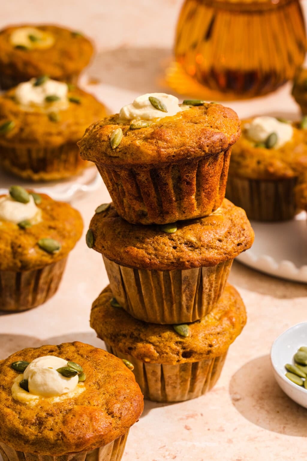 A stack of three pumpkin muffins topped with seeds and cream, surrounded by more muffins on a light surface.