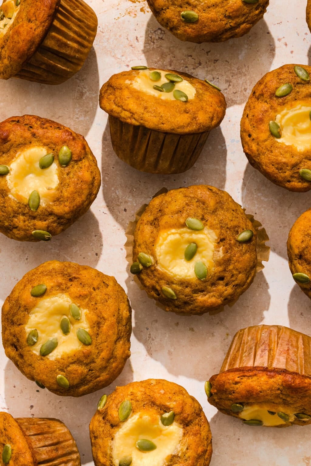 Overhead view of pumpkin muffins with cream cheese centers and green pumpkin seeds on top, arranged on a light surface.