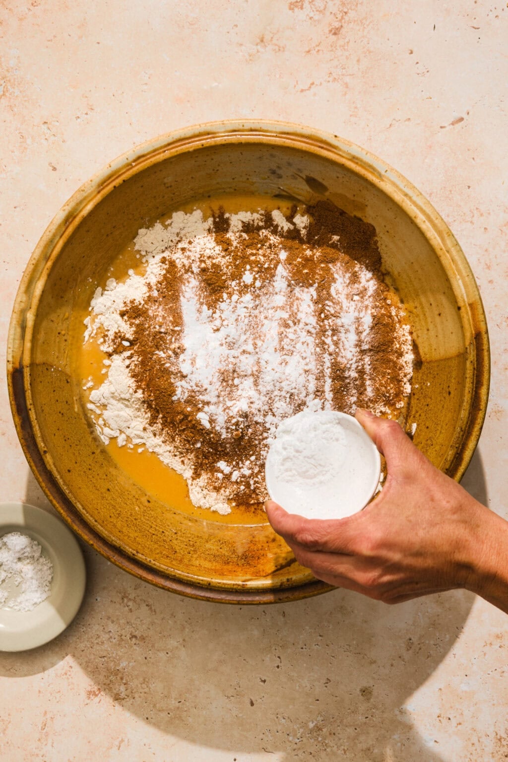A hand adds flour to a bowl containing flour, cinnamon, and other dry baking ingredients on a beige countertop.