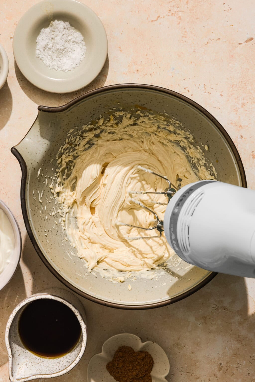 A hand mixer blends creamy batter in a bowl, surrounded by small dishes of powdered sugar, coffee, and spices on a light countertop.