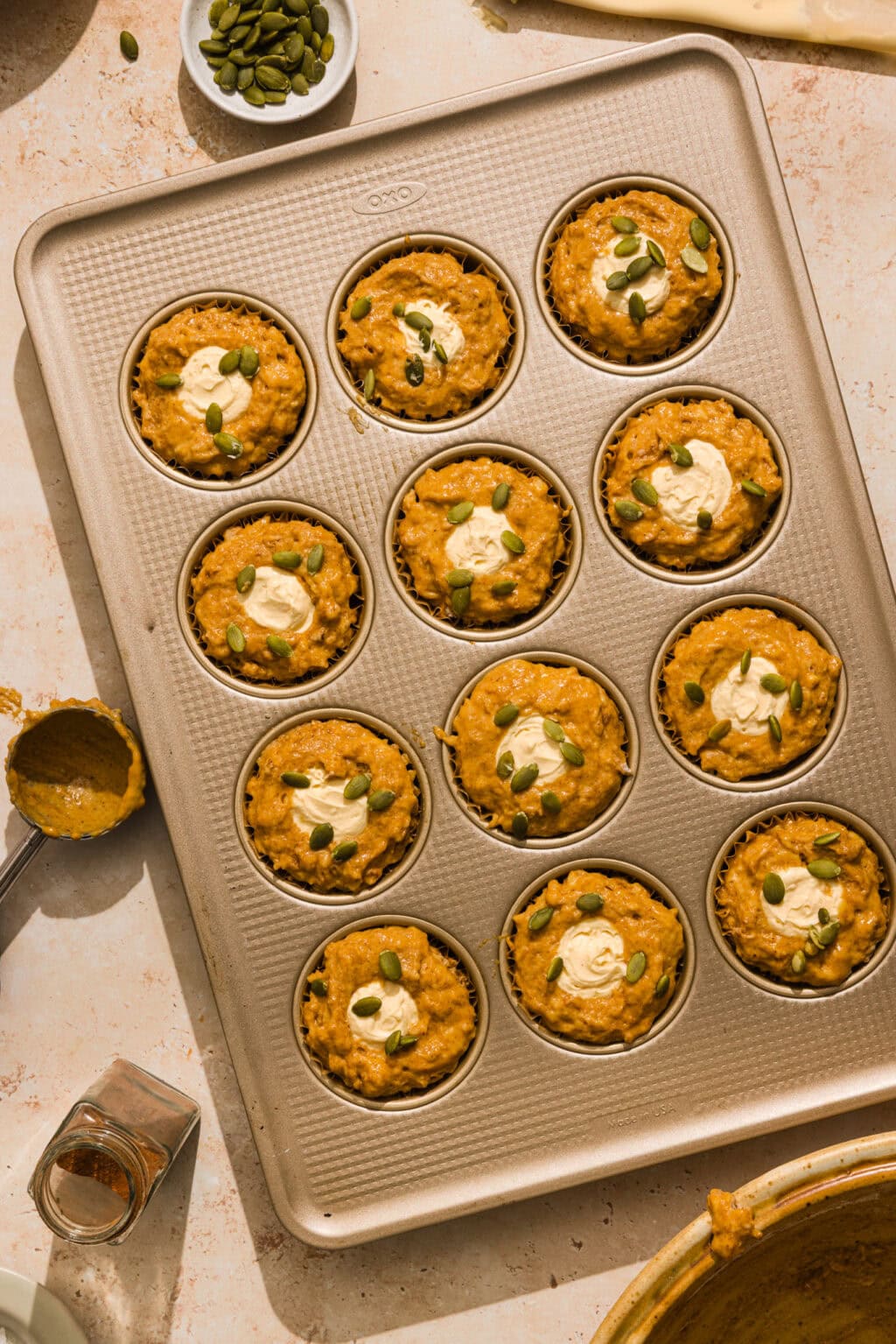 A muffin tin filled with pumpkin muffins, each topped with a dollop of cream and a sprinkle of pumpkin seeds, sits on a light countertop with baking ingredients nearby.