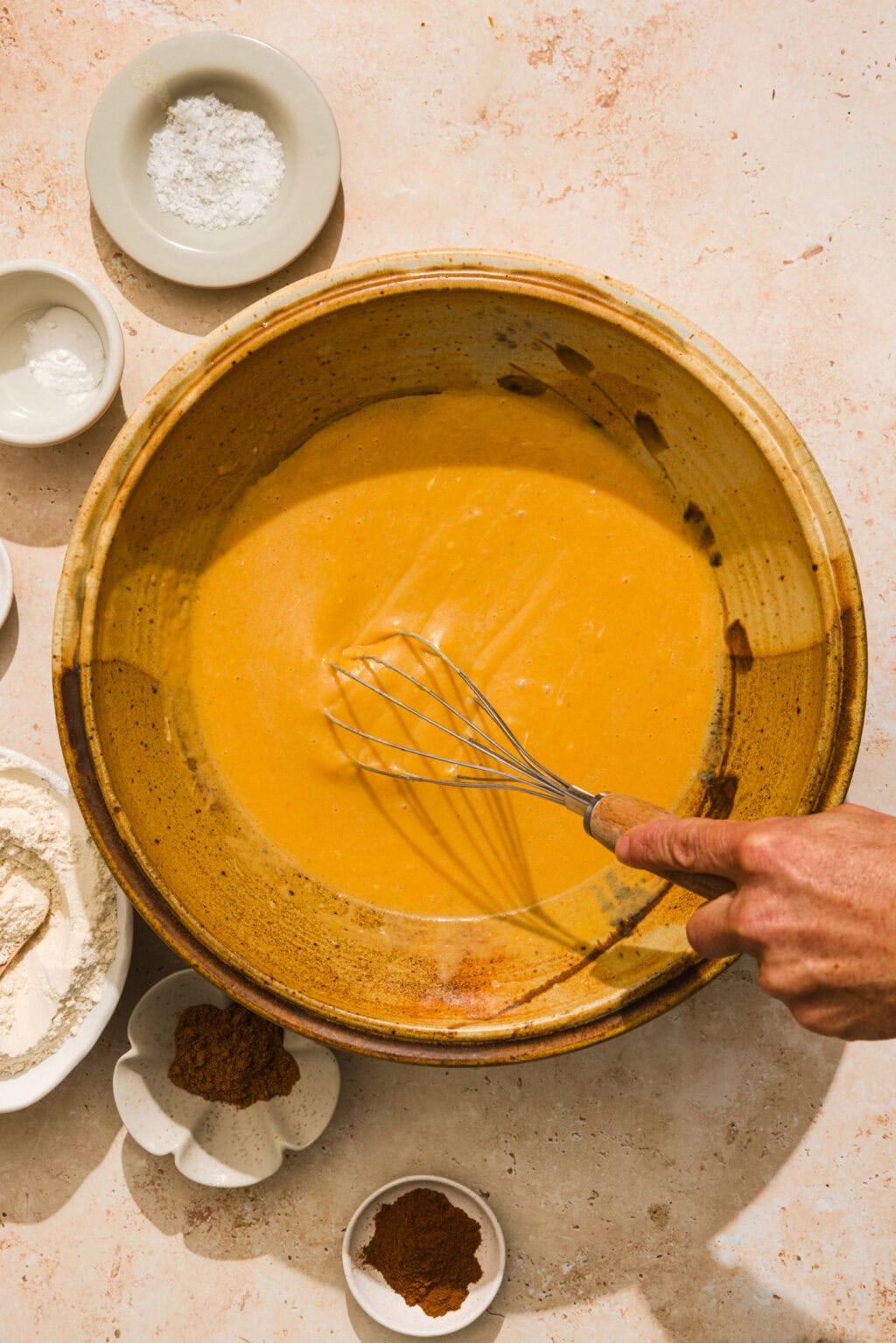 A hand whisks a smooth orange batter in a large ceramic bowl, surrounded by small bowls of flour, spices, and baking powder on a light-colored countertop.