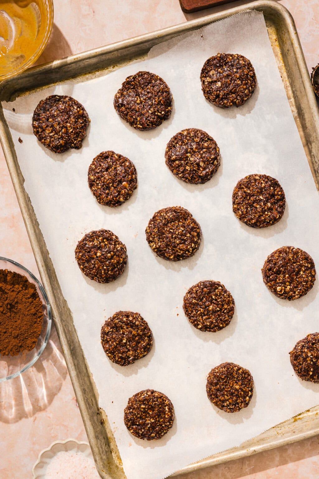 Fifteen round chocolate oat cookies are arranged evenly on a parchment-lined baking sheet. Bowls with cocoa powder and other ingredients are visible around the tray on a light pink surface.