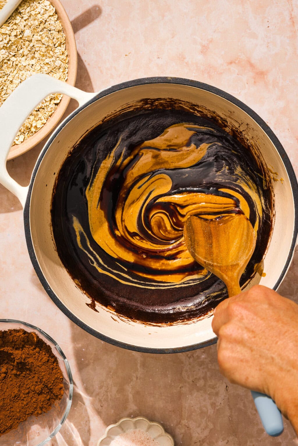 A hand stirs a mixture of chocolate and peanut butter in a pot with a wooden spoon. Nearby are bowls of oats, cocoa powder, and a small dish of salt on a pink countertop.