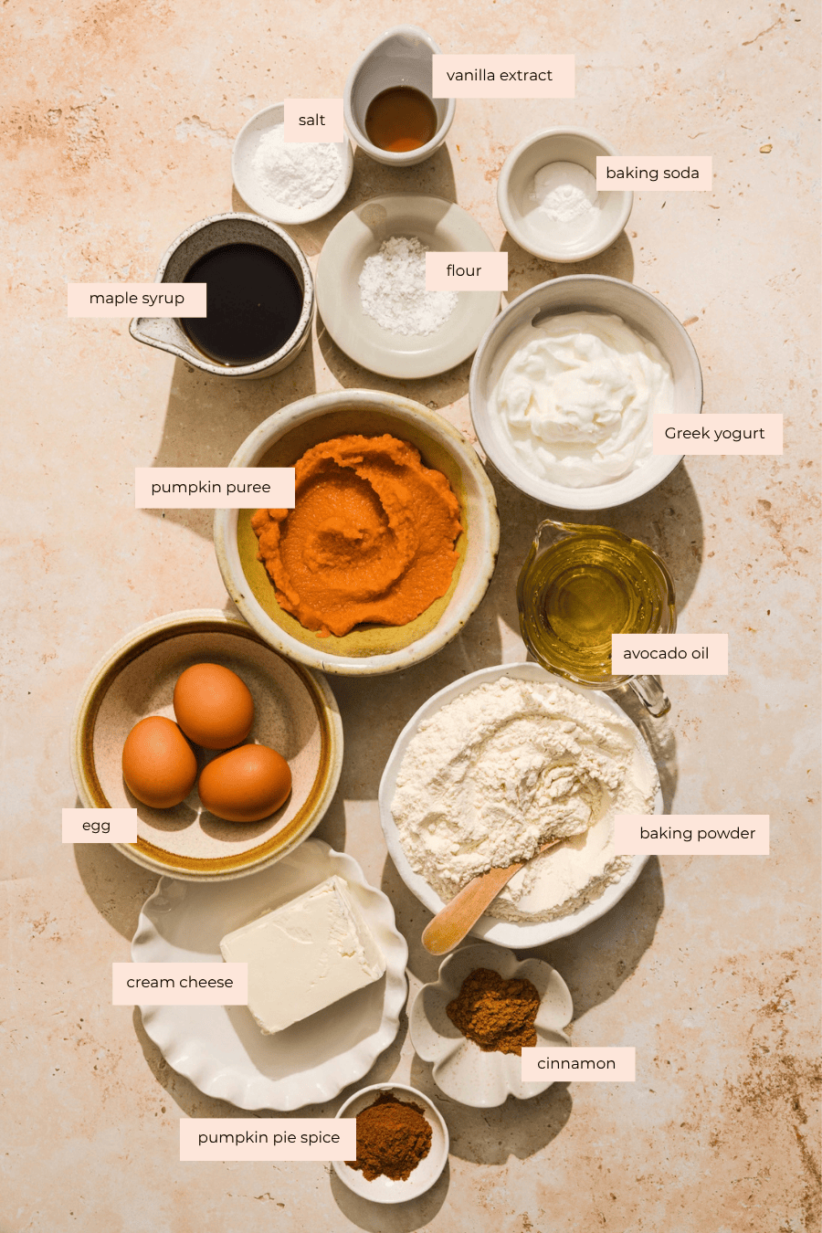 Assorted pumpkin bread ingredients for Pumpkin Cream Cheese Muffins (7g protein!) with labeled bowls of pumpkin puree, eggs, cream cheese, maple syrup, flour, baking powder, Greek yogurt, and spices.