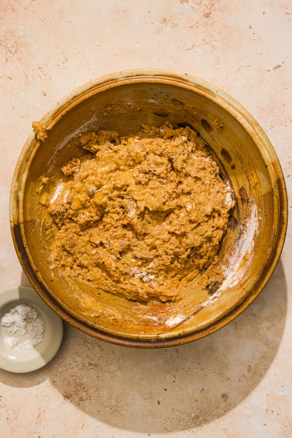 A brown ceramic bowl filled with thick, textured Pumpkin muffin batter sits on a light tan surface. Next to it is a small bowl containing a small amount of flour.